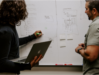 Two co-workers discuss project management, with one writing on a whiteboard and the other attentively listening.