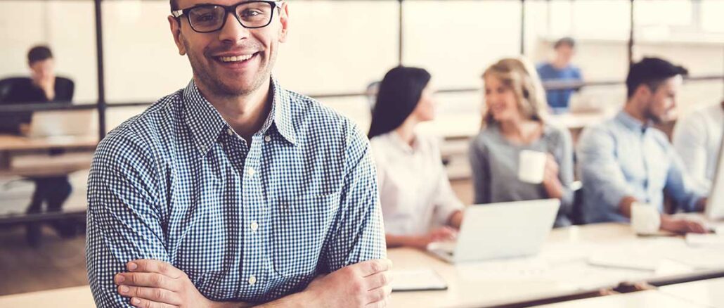 Handsome businessman is looking at camera and smiling, in the background his colleagues are working in office